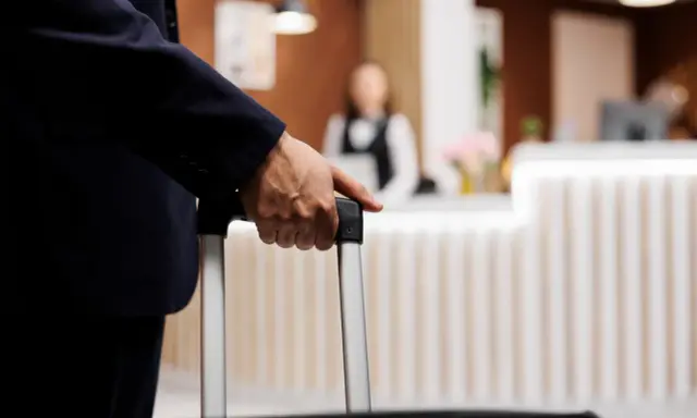 Mężczyzna w garniturze trzyma walizkę, zbliżając się do recepcji hotelowej. Czas na depozyt w hotelu i nowy początek podróży.