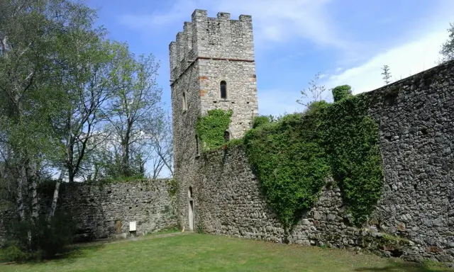 Antica torre del forte di Orino, circondata da mura in pietra e vegetazione rigogliosa, sotto un cielo azzurro.