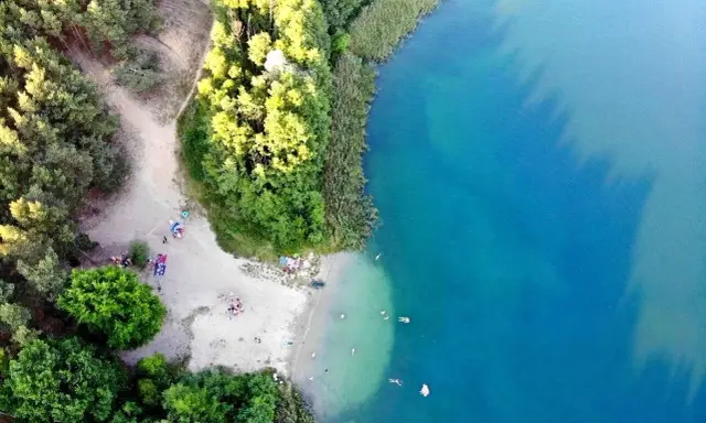 Plaża nad jeziorem tętni życiem. Ludzie odpoczywają na piasku, kąpią się w krystalicznie czystej wodzie, ciesząc się letnim dniem.