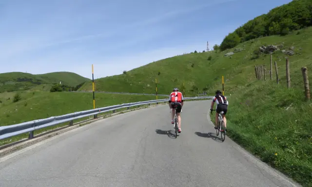 Ciclisti in salita su una strada panoramica, con verdi colline e cielo azzurro. Un'avventura che sembra un **passo cento croci**.