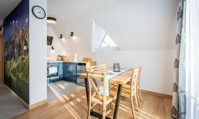 Przytulny apartament pod wierchem. W kuchni niebieskie szafki, jadalnia z drewnianym stołem. Na ścianie fototapeta z górami.