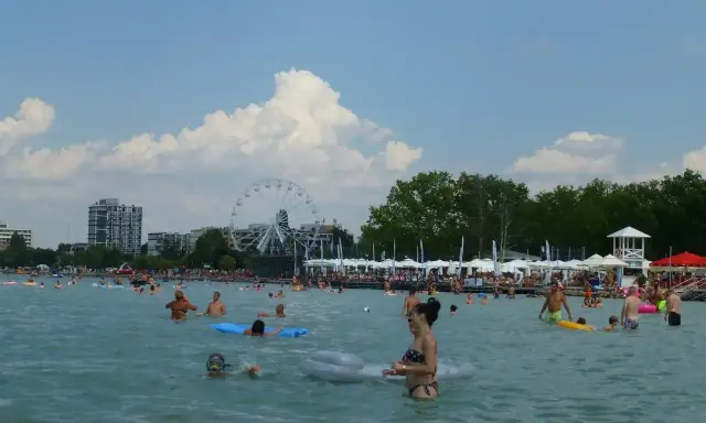 Tłum ludzi cieszy się słońcem i wodą na **Balaton plaża**. W tle widać diabelski młyn i budynki.