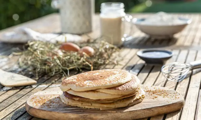 Tortitas de avena recién hechas, apiladas en una tabla de madera. Ingredientes como huevos y leche sugieren un desayuno casero.
