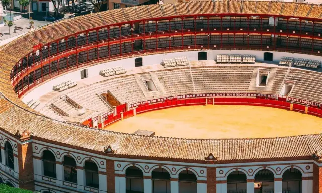 Leere Plaza de Toros mit roten Geländern und sandiger Arena. Die Sitzreihen sind leer, die Sonne scheint auf die traditionelle Architektur.