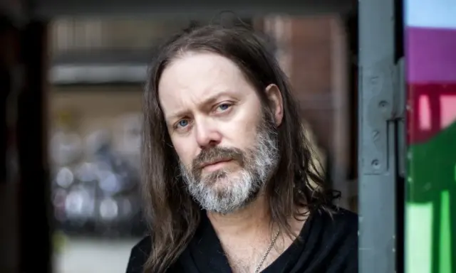 Niclas Frisk med långt hår och skägg, blickar mot kameran.
