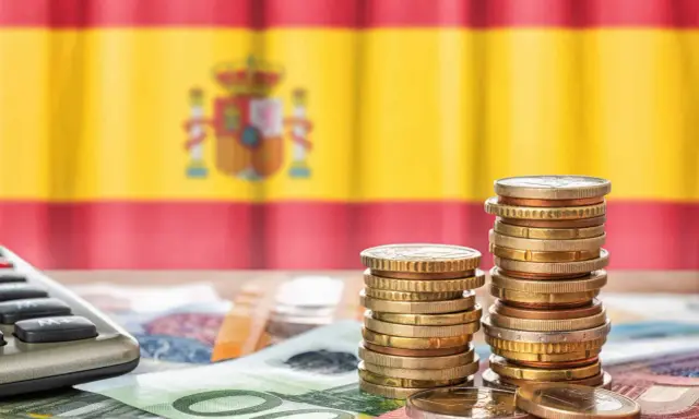 Monedas y billetes sobre una mesa, con la bandera de España de fondo. Una calculadora a la izquierda. Reflexión sobre qué son los tributos.