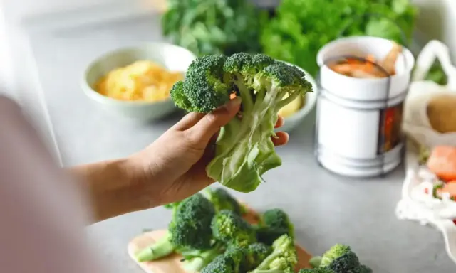 Mano sosteniendo un brócoli fresco, listo para preparar deliciosas recetas de brócoli. En el fondo, queso rallado y zanahorias.