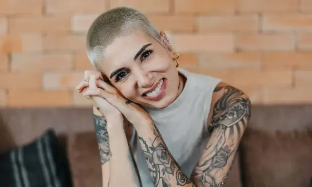 Lächelnde Frau mit Buzz Cut und vielen Tattoos, die ihre Hände vor dem Gesicht verschränkt.