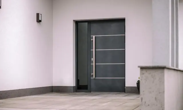 Nowoczesne drzwi wejściowe, ciemnoszare, z długą, metalową klamką i bocznym przeszkleniem. Czy to drzwi stalowe czy aluminiowe?