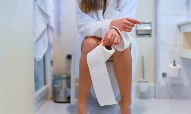 Kobieta w szlafroku siedzi na toalecie, trzymając rolkę papieru. Zastanawia się, czy jej pieniący się mocz to powód do niepokoju.