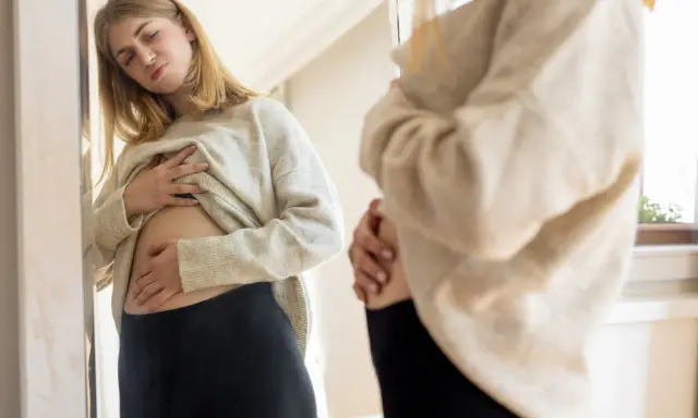 Ragazza si guarda allo specchio, toccandosi la pancia gonfia sopra i pantaloni stretti.