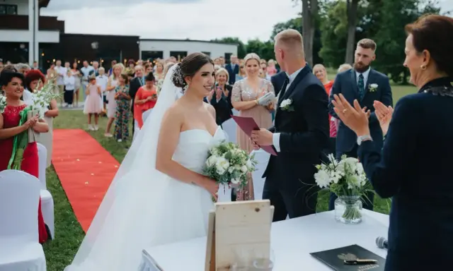Para młoda podczas ceremonii ślubu cywilnego na zewnątrz. Panna młoda w sukni, pan młody w garniturze. Goście obserwują.