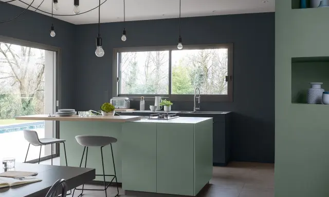 Cuisine moderne aux murs gris anthracite. L'îlot central vert sauge et le plan de travail en bois clair invitent à la convivialité. Quelle peinture pour plafond ? Un blanc mat pour agrandir l'espace.