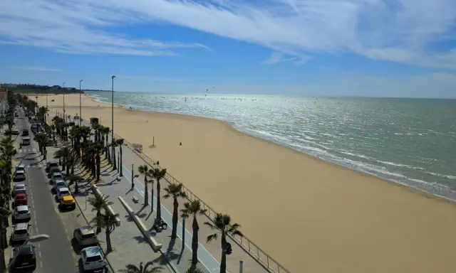 Sonniger Strand mit Palmen und Autos in Sanlúcar de Barrameda. Das Meer glitzert, am Horizont sind Kitesurfer zu sehen.