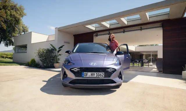 Mujer sonriente junto a un coche alto y morado, aparcado frente a una casa moderna con puertas abiertas.