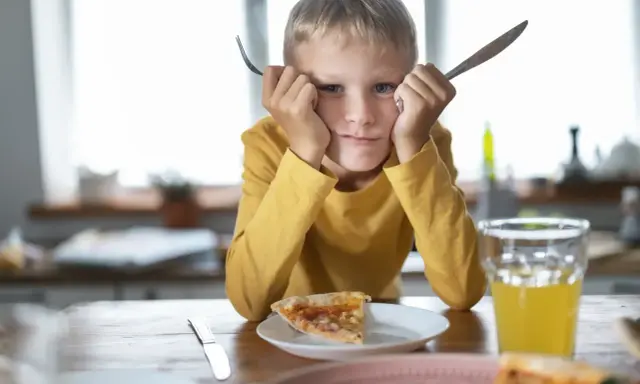 Chłopiec z widelcem i nożem w dłoniach, z miną mówiącą "dziecko nie chce jeść". Przed nim kawałek pizzy i szklanka soku.