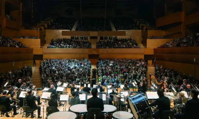 Orquesta sinfónica tocando para un público numeroso. Un director dirige la orquesta, creando una atmósfera mágica para los conciertos infantiles.