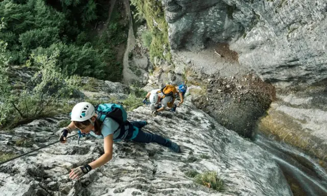 Osoby w kaskach i uprzężach pokonują skalną ścianę, używając liny i karabinków. Cały niezbędny via ferrata sprzęt zapewnia im bezpieczeństwo podczas wspinaczki.