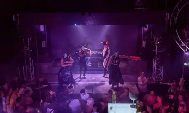 Performers in masks and elaborate outfits dance on stage at one of Valencia's vibrant gay bars, while a crowd watches below.
