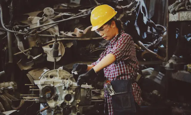 Mujer con casco amarillo y gafas de seguridad trabajando en un motor. Usa guantes negros y un delantal de mezclilla, demostrando el uso de los equipos de protección individual.