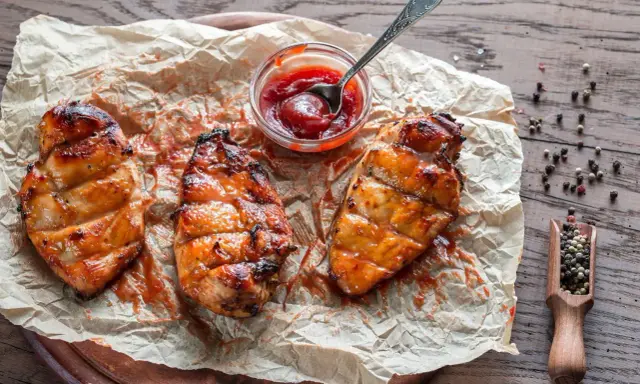 Saftiges Grillfleisch zart machen: Drei gegrillte Hähnchenbrustfilets mit Sauce auf Backpapier, daneben eine Schale Ketchup und eine Holzschaufel mit Pfefferkörnern.