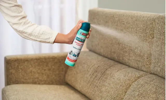 Mano rociando desinfectante en spray sobre un sillón de tela. Una forma fácil de saber como limpiar sillones de tela y eliminar olores.