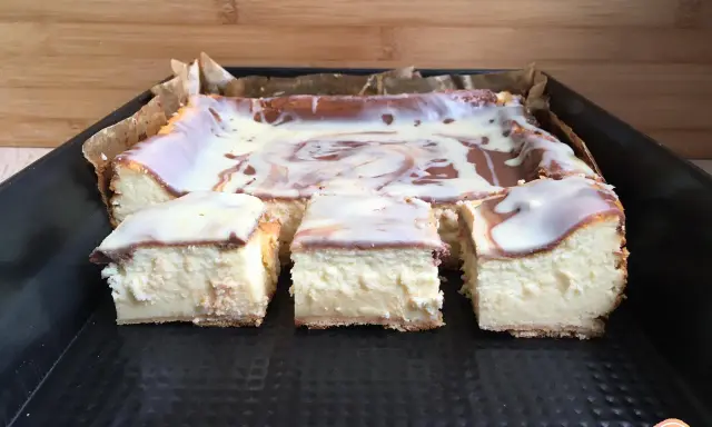 Pyszny sernik z mlekiem, pokrojony na kwadraty, polany białą polewą z czekoladowymi wzorami.