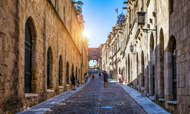 Sonnenbeschienene Gasse in Rhodos Stadt mit Kopfsteinpflaster und historischen Gebäuden. Touristen schlendern durch die mittelalterliche Stadt.