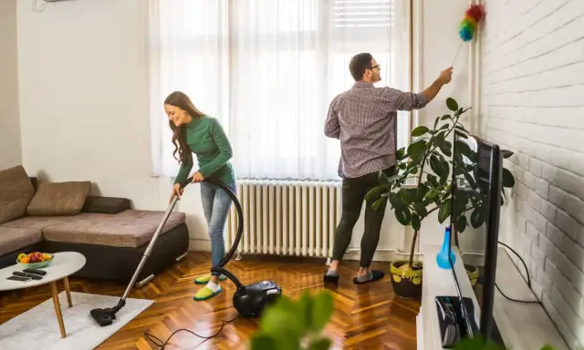 Pareja colaborando en como limpiar la casa: ella aspira el suelo y él quita el polvo de las paredes.
