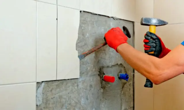 Man con guantes rojos usa un cincel y martillo para quitar azulejos y cemento de una pared.