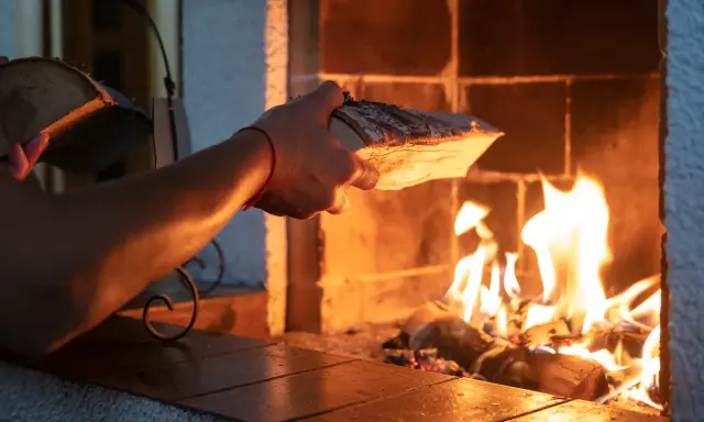 En hand lägger ved i en sprakande brasa. Det syns tecken på soteld i eldstaden.
