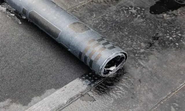 Rolka papy na zimno leży na mokrym dachu. Widać fragmenty czarnej papy i charakterystyczny wzór na rolce.