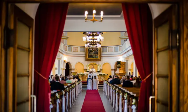 W kościele trwa ślub jednostronny. Panna młoda i pan młody stoją przed ołtarzem, a ksiądz udziela im błogosławieństwa.