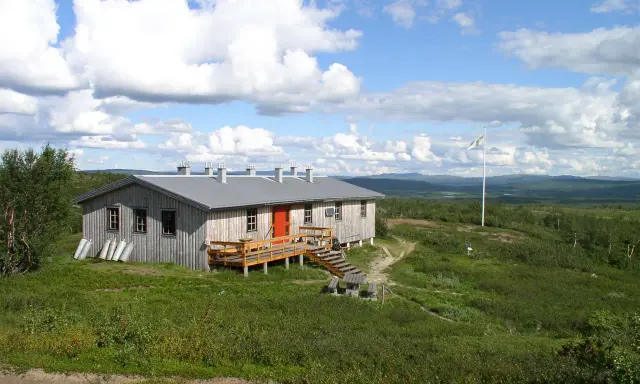 Kungsleden stugor i fjällandskap med molnig himmel. Enkel träbyggnad med veranda och flaggstång.