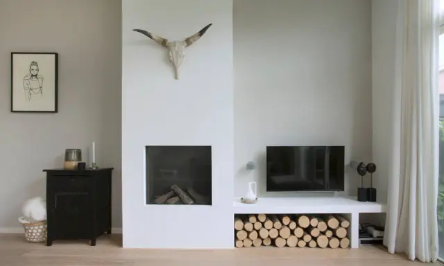 Un salon moderne avec une cheminée blanche épurée, un crâne de taureau, du bois de chauffage et une télévision. Un avant-après réussi pour relooker une cheminée.