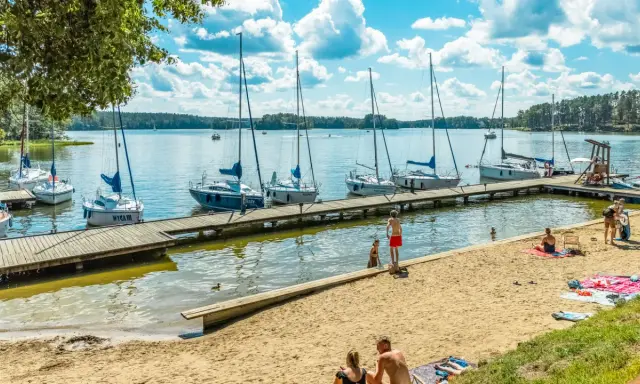 Słoneczny dzień nad jeziorami mazurskimi. Drewniane molo z żaglówkami, piaszczysta plaża z wypoczywającymi ludźmi i błękitne niebo z chmurami.