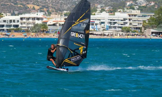 Ein Mann beim Windsurfen in Griechenland, mit Blick auf eine belebte Küste und Berge im Hintergrund.