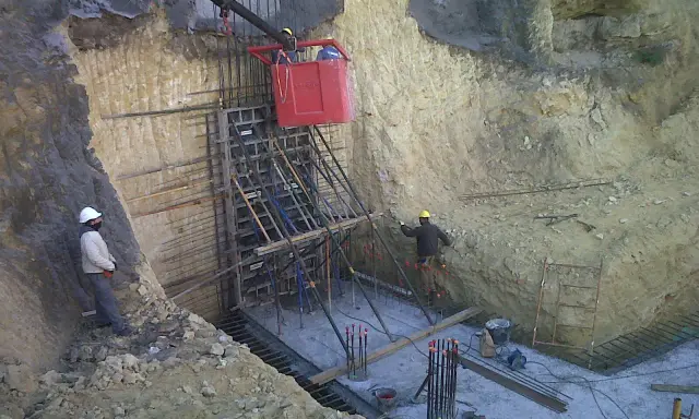 Trabajadores instalan encofrado para muro pantalla, con bataches y estructura de acero en obra.