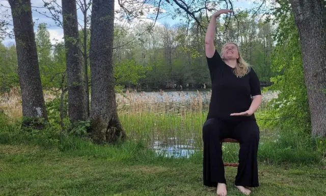 Kvinna i svart klädsel utför yoga på en stol vid en sjö. Naturen är grönskande med träd och vass.