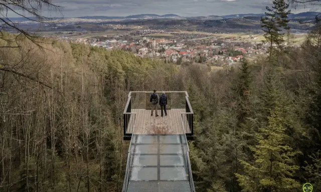 Dwie osoby stoją na szklanym tarasie widokowym, podziwiając panoramę Starego Sącza i okolic.