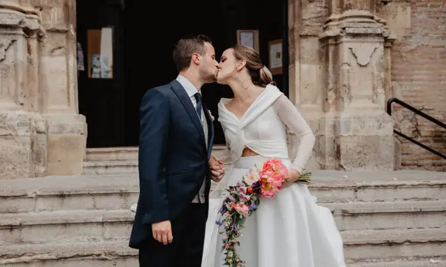 Novios besándose frente a una iglesia, ella lleva un ramo de flores. Un momento especial tras las lecturas de boda religiosa.