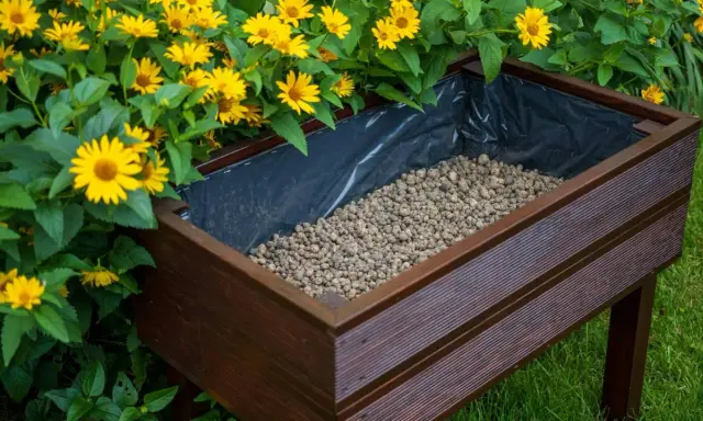 Wysoka donica z brązowego drewna, wyłożona czarną folią, wypełniona żwirem. Obok kwitną żółte kwiaty. Idealne rozwiązanie, co zamiast keramzytu do doniczki.