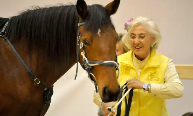 Linda Tellington mit einem braunen Pferd, sie trägt eine gelbe Weste und eine Blume im Haar.
