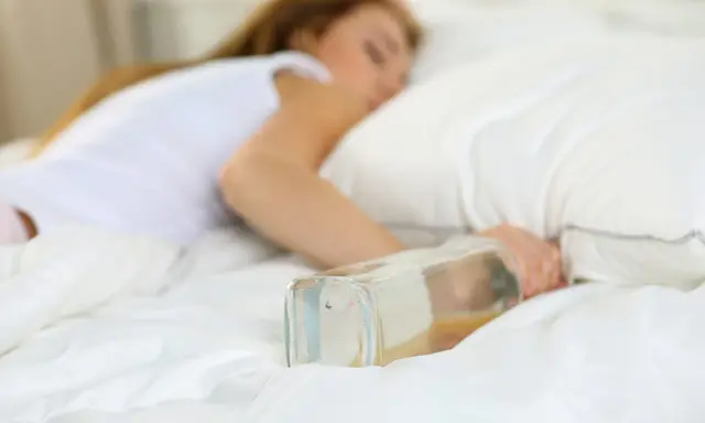 Kobieta śpi w łóżku z butelką alkoholu. Problemy ze snem mogą być związane z neuropatią alkoholową.