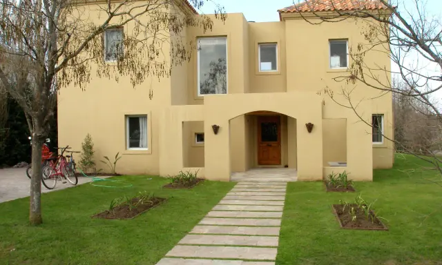 Una encantadora casa color arena con techo de tejas rojas, un camino de losas y dos bicicletas aparcadas en el jardín.