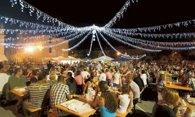 Festa notturna con migliaia di persone sedute ai tavoli, illuminate da festose luminarie. Si celebra la sagra te lu purpu, un evento conviviale.
