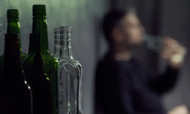 Puste butelki na pierwszym planie, w tle niewyraźna postać pijąca alkohol. Gdzie jest bezpieczna ilość alkoholu?