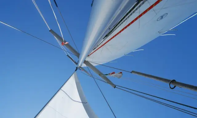 Vue plongeante sur le mât et les voiles d'un voilier, le ciel bleu intense en arrière-plan. L'image capture l'essence de la navigation, le vent gonflant les voiles.