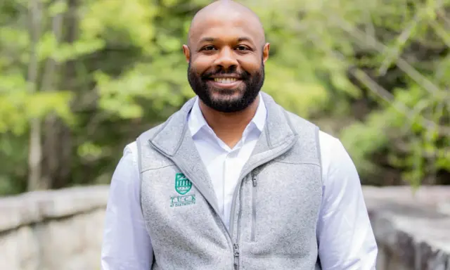 A smiling man with a beard wears a grey vest over a white shirt. The vest has a Tuck at Dartmouth logo, suggesting a curated professional image.