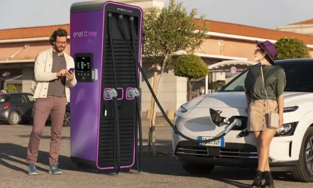 Coppia in una stazione di ricarica Enel X Way. Lei attende mentre l'auto si carica, lui controlla lo smartphone. Ottime recensioni per questa stazione di ricarica.